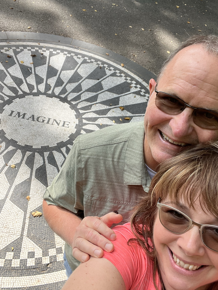 Smiling couple takes selfie next to the Imagine mosaic in Central Park, symbolizing new beginnings and possibilities.