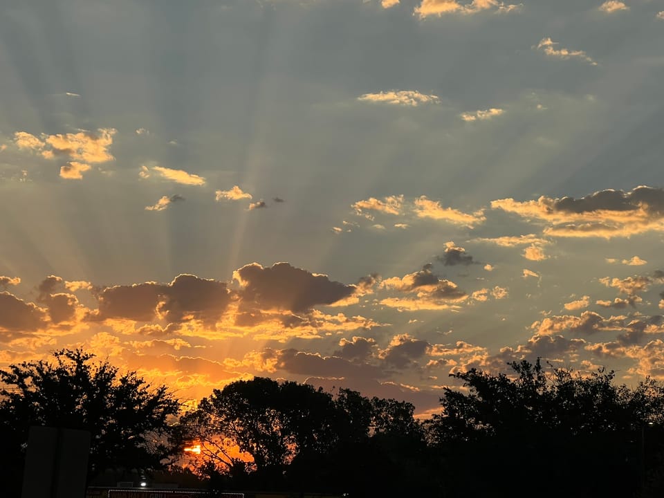 Dramatic sunset with golden rays streaming through clouds above silhouetted trees, captured during evening walk.